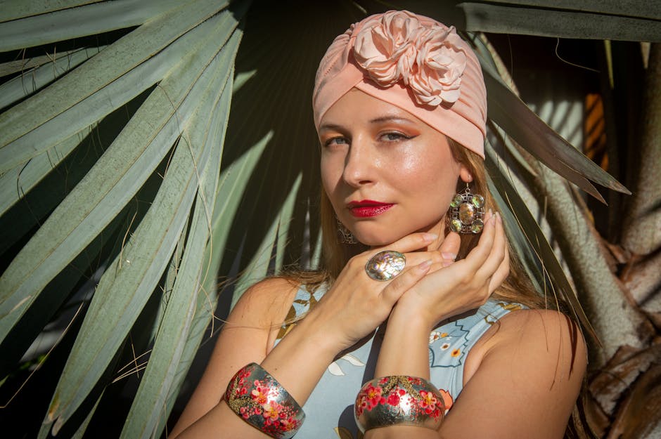 A stylish woman in floral headwear poses elegantly with bold jewelry against a leafy backdrop.