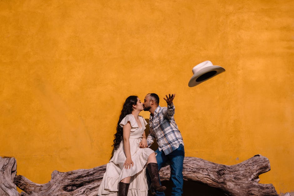 Romantic moment of a couple kissing while sitting on a log, highlighted by a vivid yellow background and a playful thrown hat.