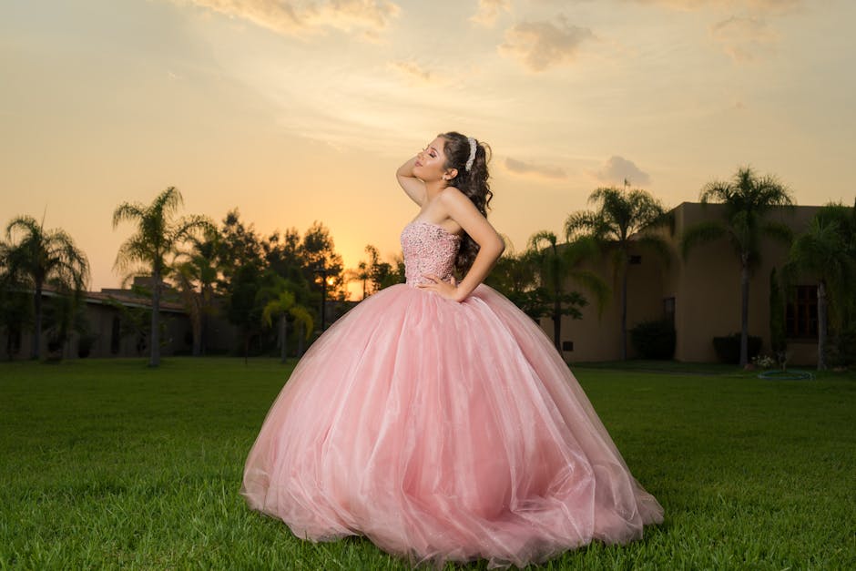 A beautiful woman in a glamorous pink gown poses elegantly in a garden at sunset.