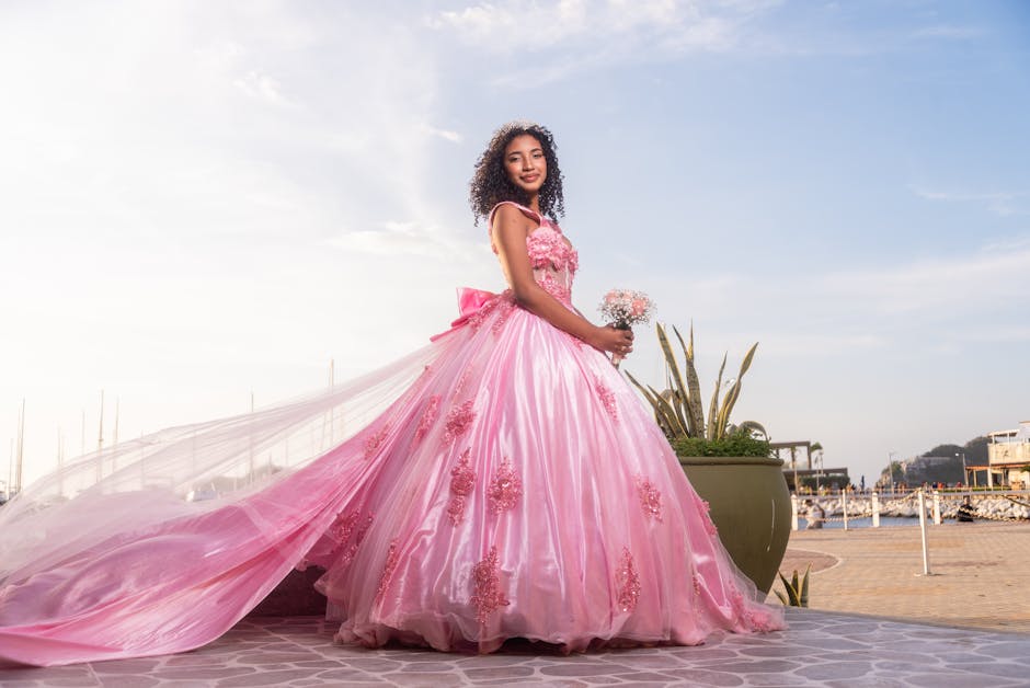 A young woman in a flowing pink gown poses by a marina, holding flowers under a sunny sky.