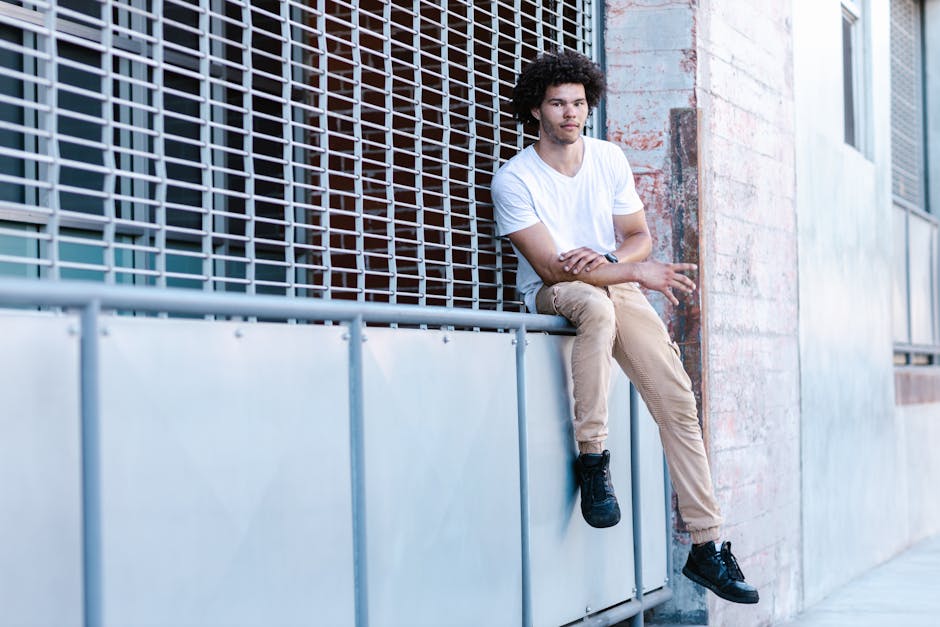 A young man in casual attire sits on a metal railing in an urban setting, showcasing modern street fashion.