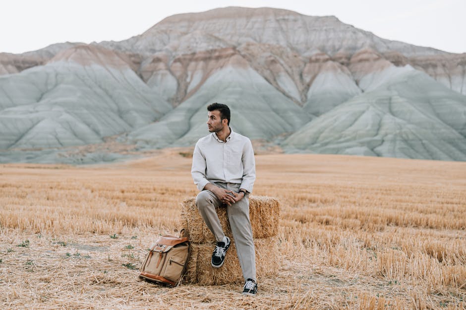 Stylish man sitting on a hay bale in the scenic fields of Nallıhan, Turkey, with picturesque mountains in the background.