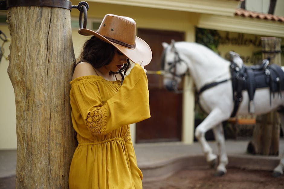 Woman in yellow dress and cowboy hat standing by a tree with a white horse in the background.